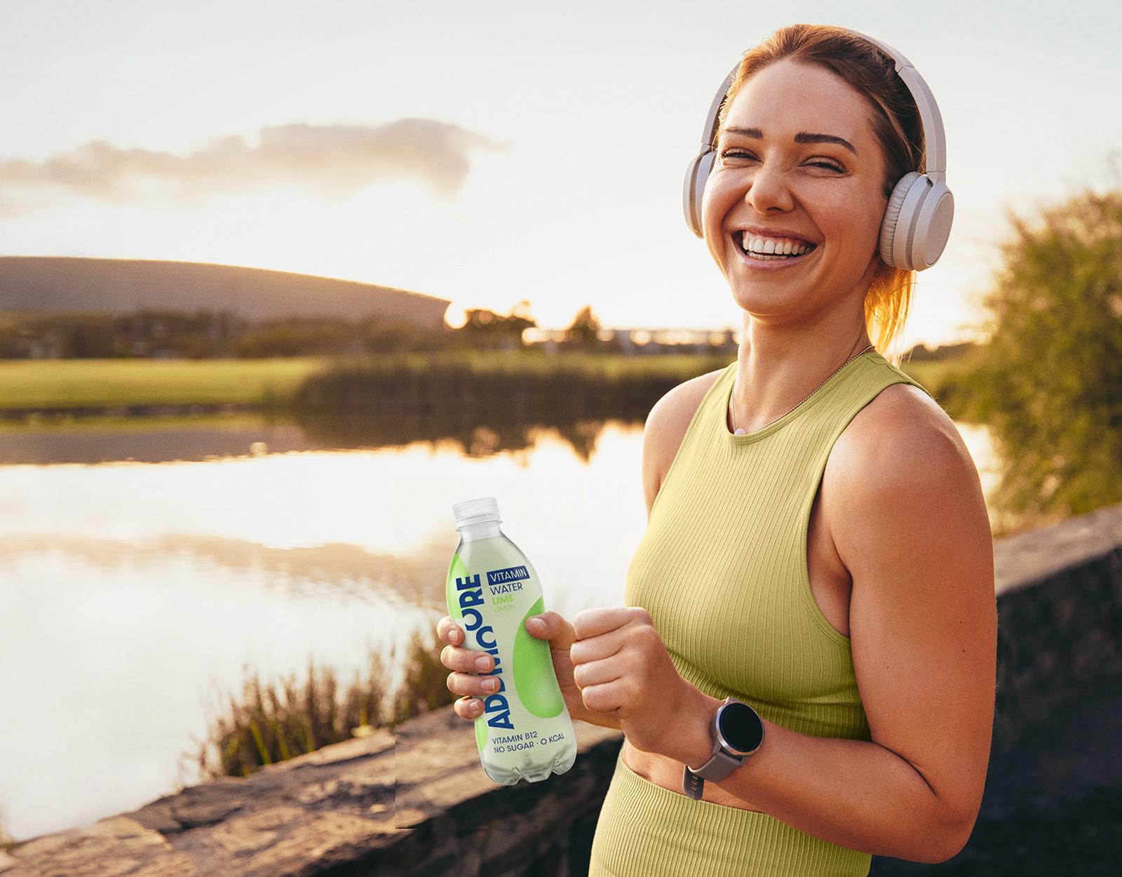Lachende vrouw met koptelefoon bij een meer in avondlicht houdt een fles Add Mooore Vitamin Water vast